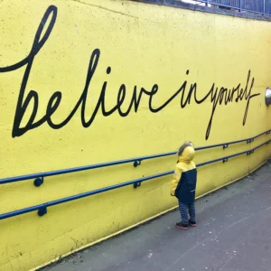 Image shows child with a coat on facing a large mural that says 'Believe in Yourself'