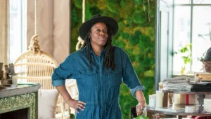 Image shows woman leaning on a surface inside her studio. She is wearing a wide-brimmed black hat, and a blue-jean jumpsuit.
