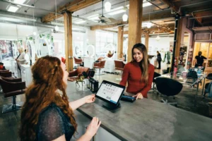 Image shows two people performing a transaction at the front desk of a salon. One person is sitting behind the front desk with an ipad in front of her. The other person is smiling and paying for her services. The background of the image is a brightly lit salon.