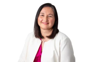 Image shows woman, with short black hair, smiling, in front of a white backdrop. She is wearing a white blazer and a has a bright pink, shiny, blouse underneath.