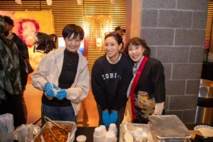 Image shows three Asian women smiling while serving food at an event.