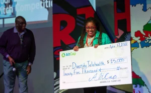 Image of woman and man on stage. Woman is smiling and holding a large check written to her business for $25,000