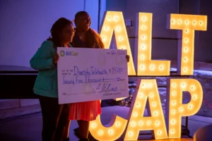 Image shows two women holding a large check. They are dimly lit by large marquee letters that spell out 'AltCap'