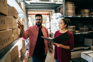 Image shows and man and a woman in a small warehouse. The man is pointing at shelves of stacked boxes while the woman holds a black binder with papers inside.