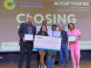 Group of 4 men and 2 women holding a large check and certificates of awards on a stage during an event