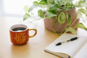 Image shows a desk with a plant, a full mug of coffee, and an open notebook with a pen on top of a blank sheet.