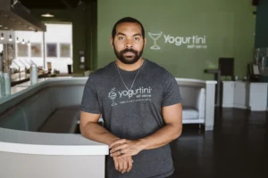 Image shows man inside a closed frozen yogurt shop. He is leaning on a counter, wearing a shirt with the logo of the business on it.