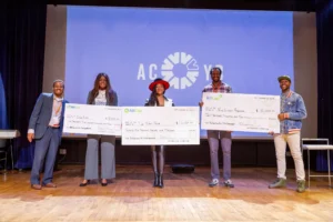 Image shows three men and two women standing on a stage in front of a large presentation screen. They are holding three large checks and smiling.