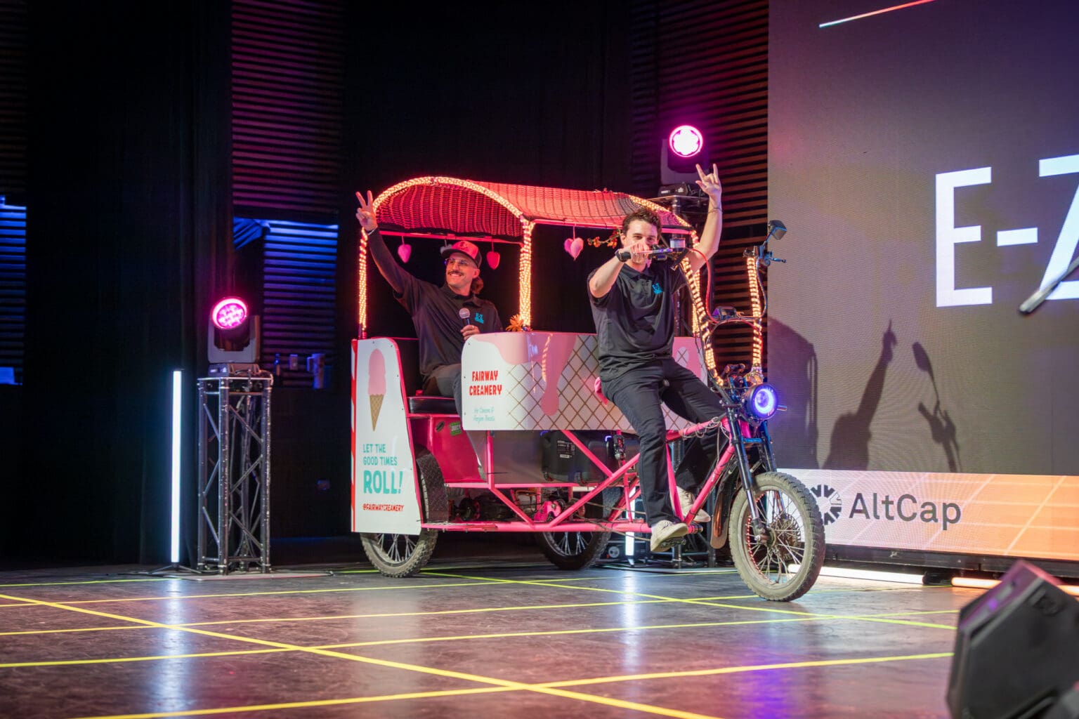 E-Z Pedicabs co-founders Atticus Sloan and Mark Manning ride a pedicab on stage at the 2025 AltCap Your Biz Pitch Competition